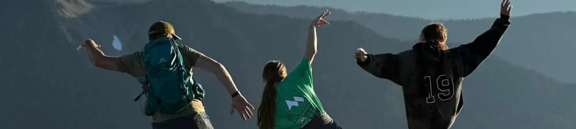 three people with their backs to the camera, striking funny poses in front of a mountain range