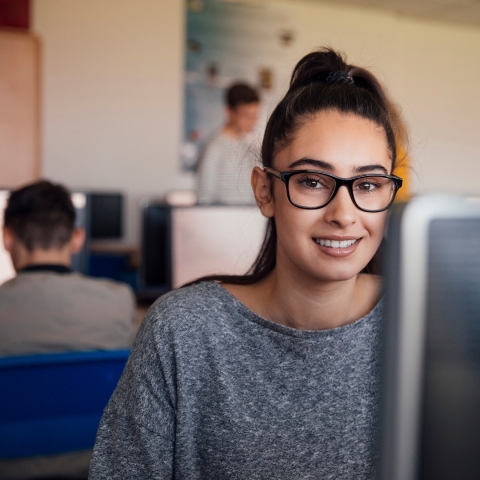Woman with dark hair and glasses sitting at a computer in a classroom