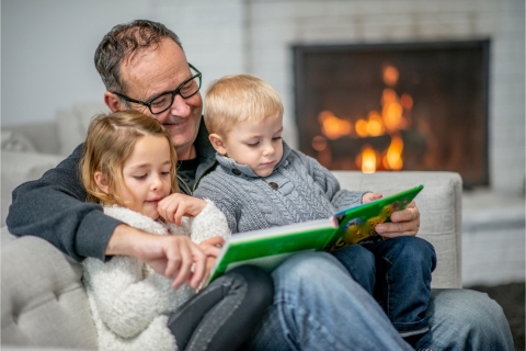 Grandpa reading a book to grandchildren next to a fireplace