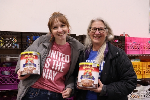 Two women in a food bank holding large cans of refried beans