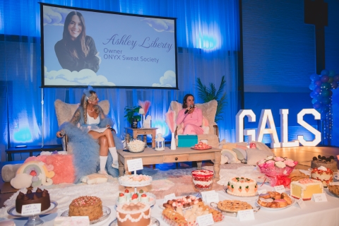 Two women speaking together on a stage with a table of desserts in front of them
