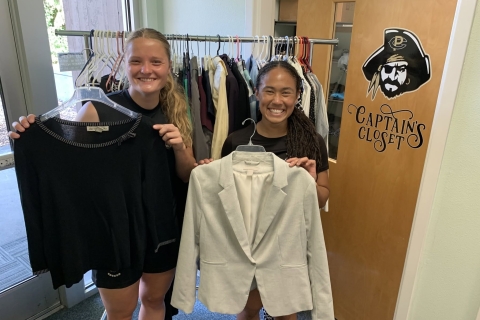 2 college students standing in front of a rack of clothing holding up work attire